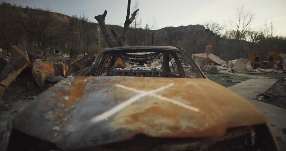 A burnt out and rusted car sits with a white x marked on the hood