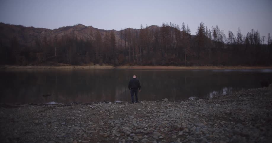 A person stands at the edge of a lake with a tree line and hills visible at dusk