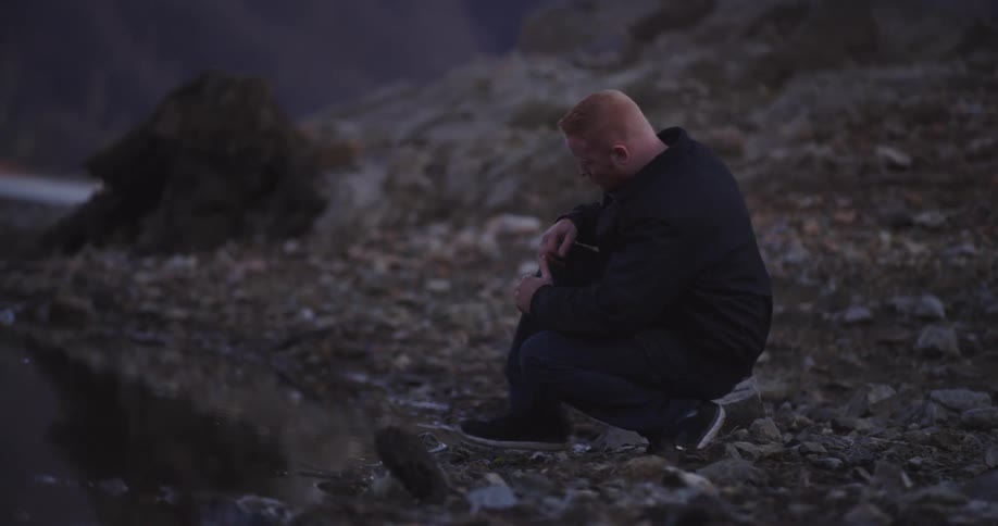 A young man bends down at the edge of a river
