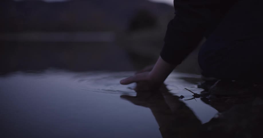 A man knees next to a river and dips his hands in the water