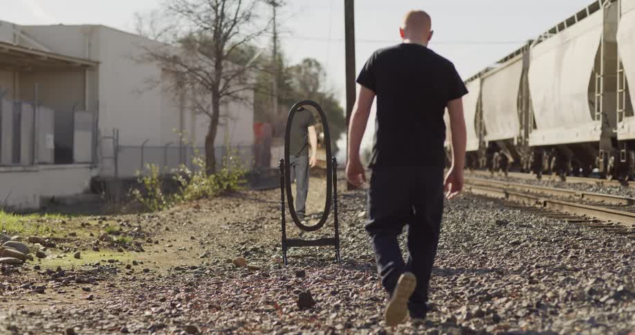 Man walk toward mirror set outdoors near passing train and warehouse