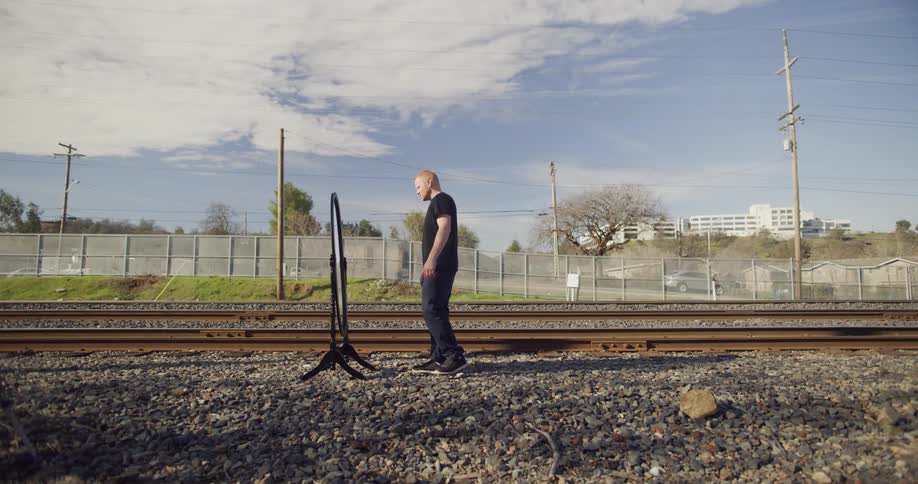 Man walks up to a mirror beside rail road tracks and reaches there are cars driving down a road behind a fence and a building on a sunny day