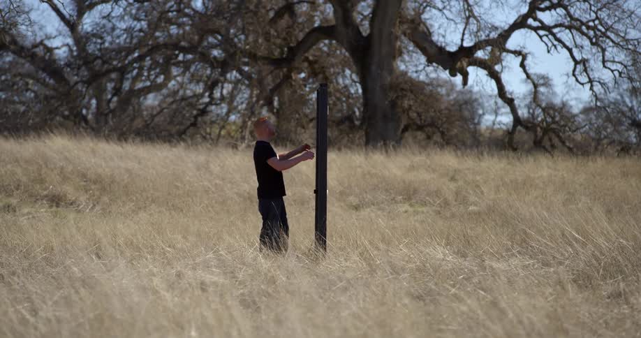 Man in black walks up to a pole in a field with tall grass and shakes it in front of trees and a blue sky on sunny day