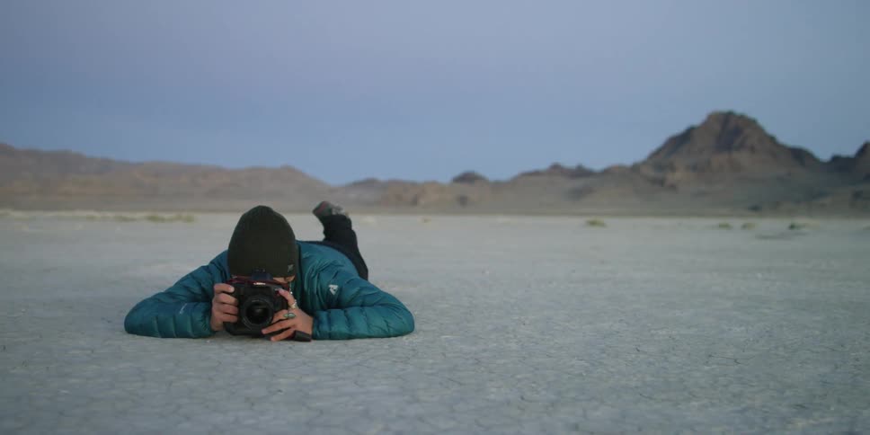 A person is lying on the ground of a desert area taking photographs