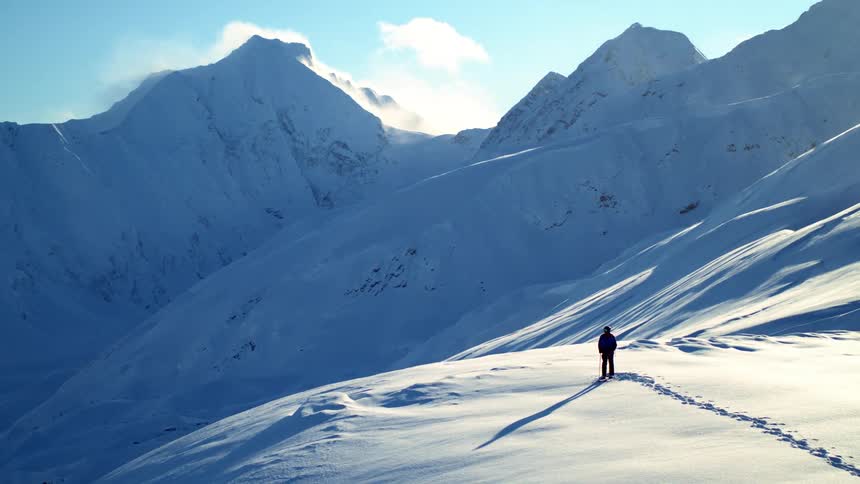 Person standing at the top of a mountain completely covered in snow next to a cliff and mountain peeks at day time