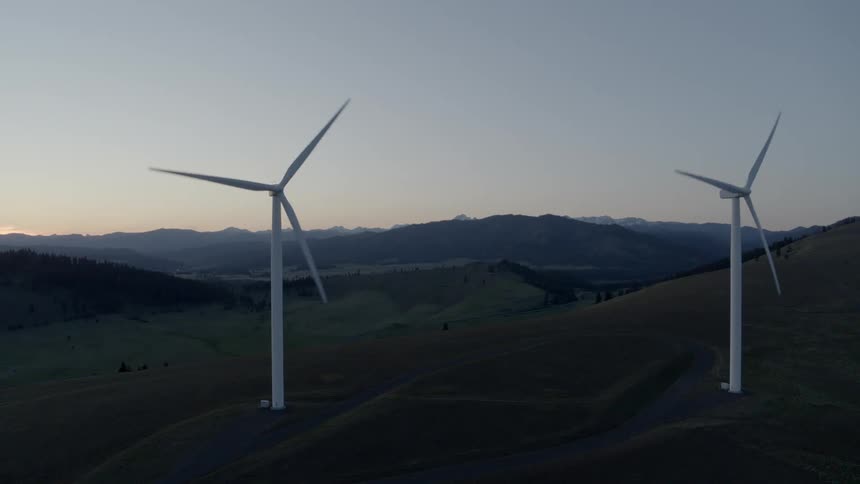 Sunset orbit around two wind turbines on rolling hills with mountains in the distance