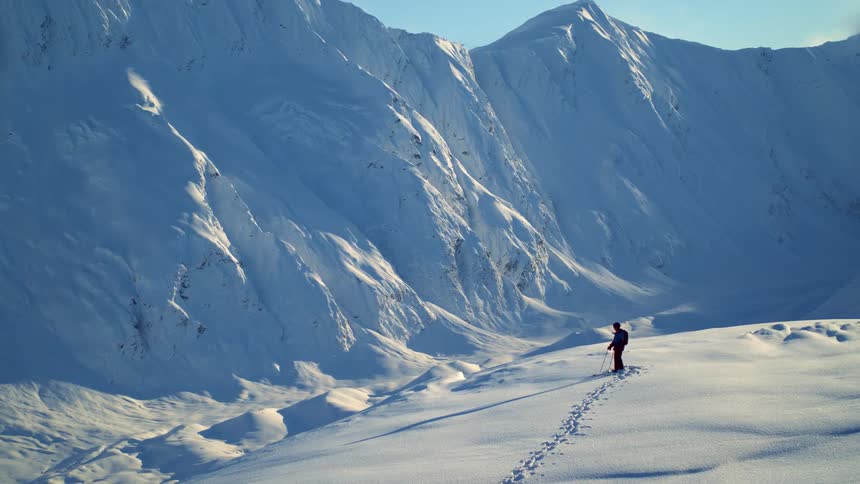 A man leaves footprints in the snow as he hikes to the end of a snow covered plateau and looks up at a mountain