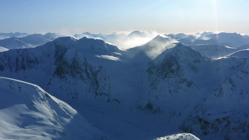 Fog swirls around the summits of a snow covered mountain range during the day