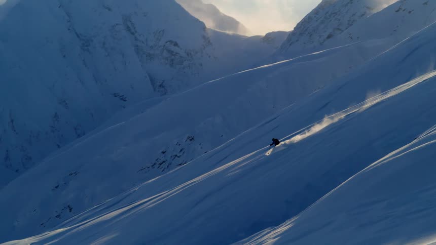 Person skis down snow covered mountain with the sun shines through the mountain peaks in the background