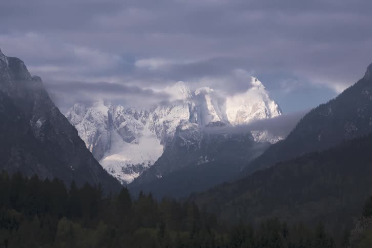 Wide shot of a snow covered mountain as clouds roll by it in the sky two smaller mountains without snow but covered in trees
