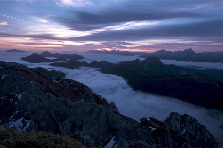 Thick mist flows like a river between the peaks of a mountain range on a cloudy evening