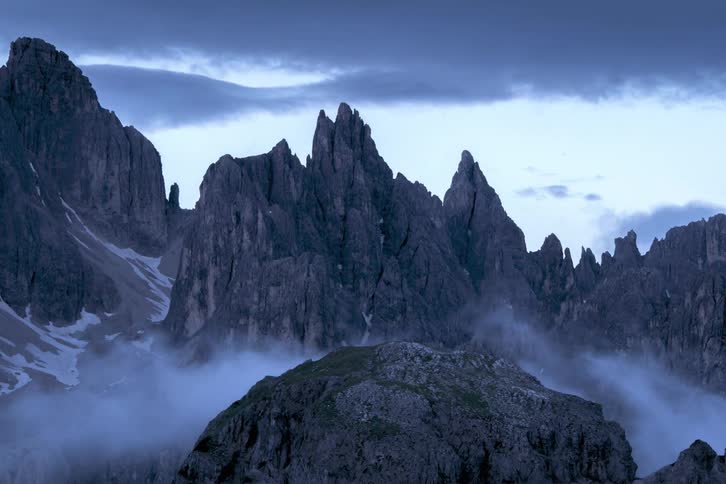 Mist flows over rocks in mountain valley with jagged cliffs and mountains rising above at night