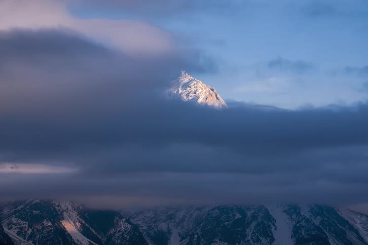 A snowy mountain peak looming over a mountain range is slowly obscured by the formation of dark clouds in the daytime sky