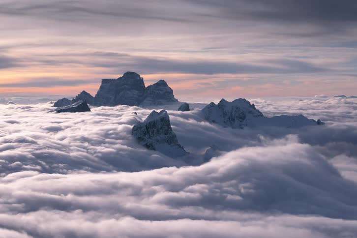 Clouds pouring over the tops of mountains at day time next to a sunset sky