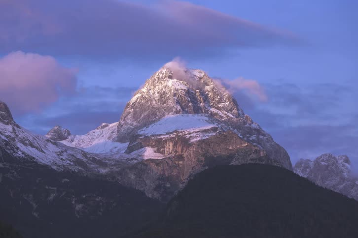 Clouds moving across the top of a snowy mountain peak at sunset