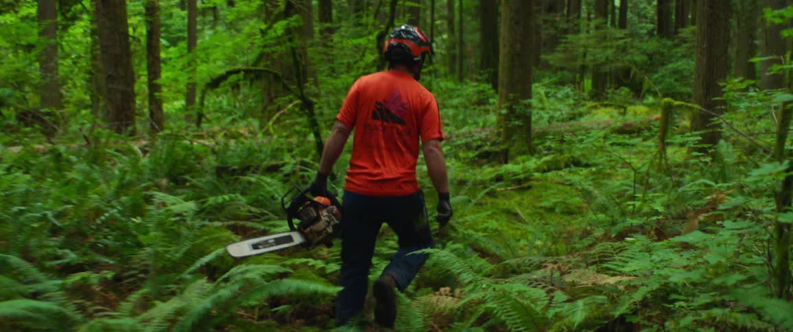 Man with a helmet and an orange shirt carries a chainsaw and walks in the woods