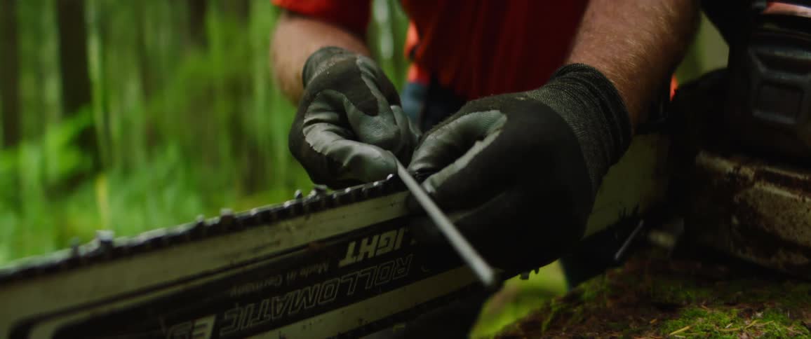Man in forest area wearing gloves sharpens a chainsaw blade with a file on a down tree log