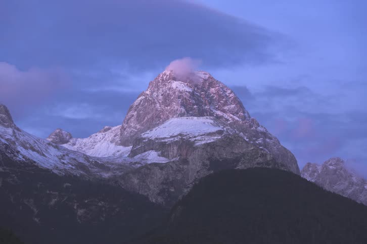 Clouds moving across the top of a mountain summit covered in light amounts of white snow at sunset