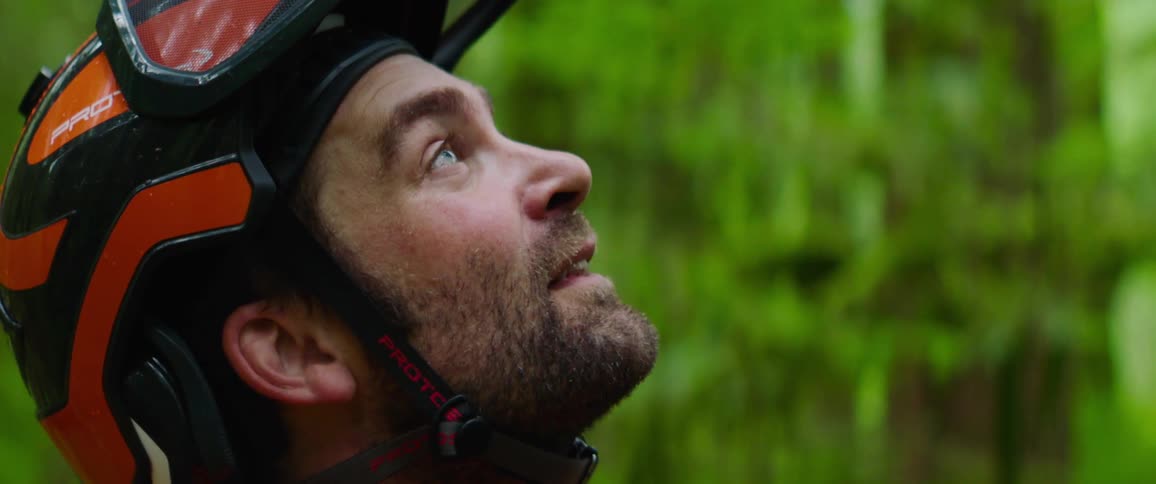 A man wearing a helmet and standing in the woods looks up and smiles