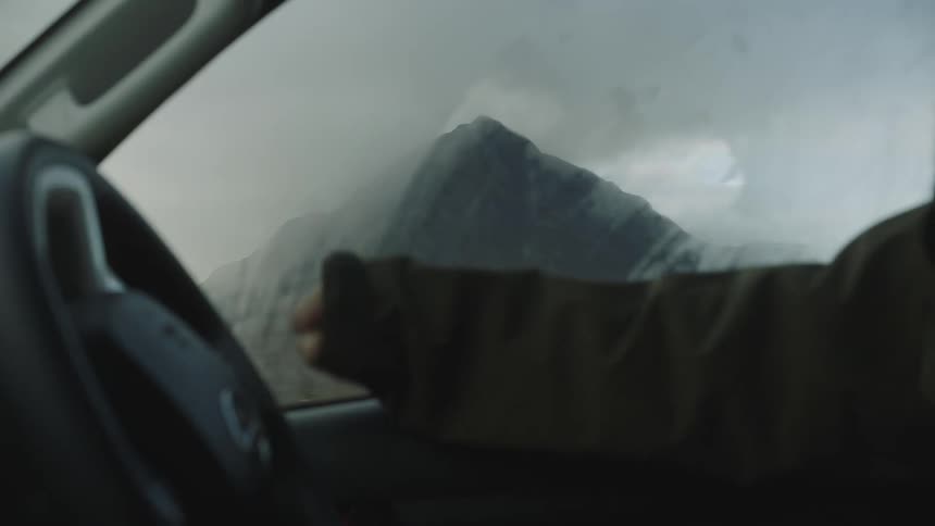 Person sits inside a car and uses the jacket sleeve to wipe off fog from the interior window to reveal a mountain