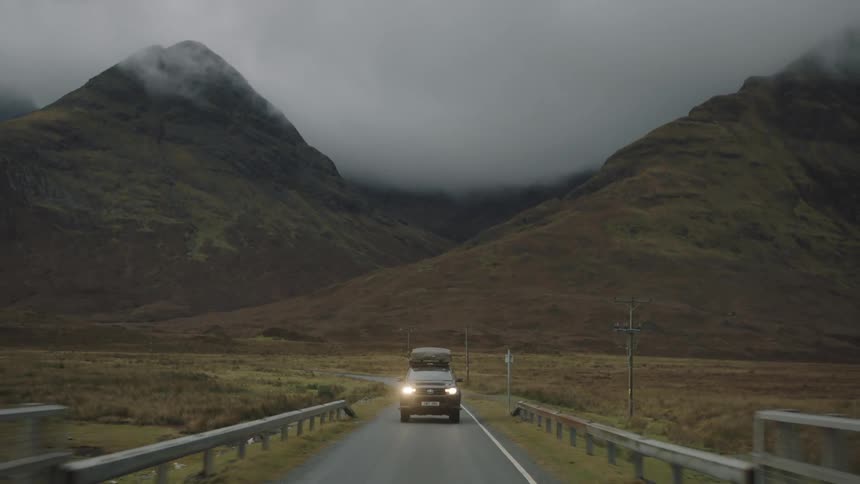 Black car with luggage on top driving down a highway in front of mountains in a remote location on a foggy day