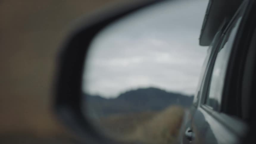 Rear view mirror of a car driving down a road with mountains in the distance at daytime