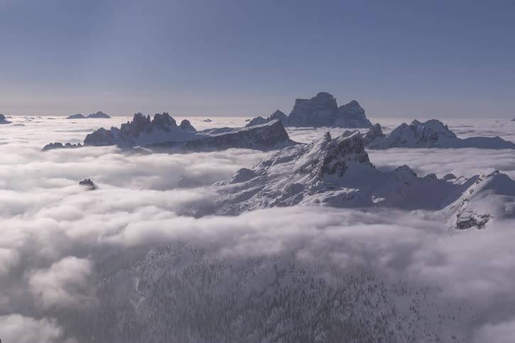 Snowy mountain peaks poke through a sea of clouds swirling above a mountain range during the day