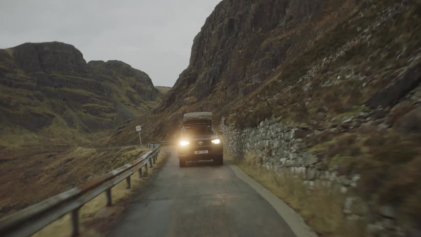 A vehicle travels along a narrow road through the mountains on a cloudy day