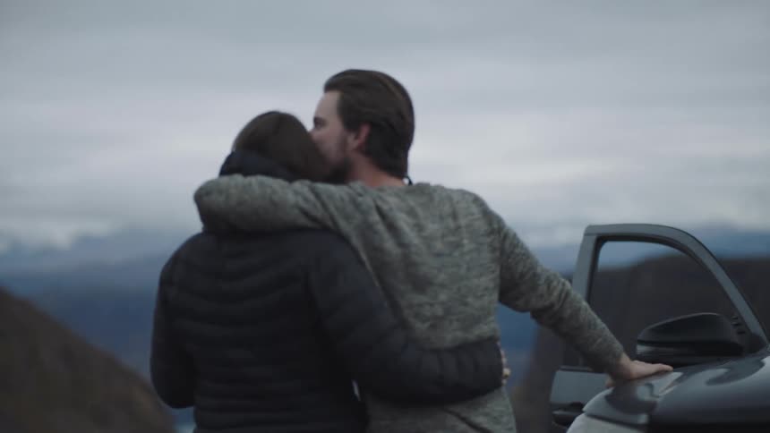 Couple standing next to a car at day time hugging and staring off at a mountain range