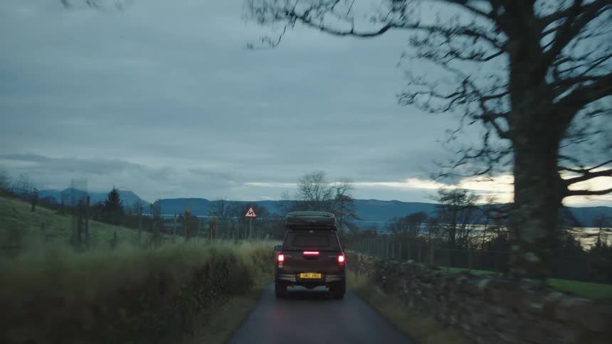 Car driving down a road at sunset in the country side in between a rock wall and grass slope