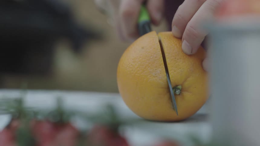 A person is holding a knife and cutting an orange in half on a white table