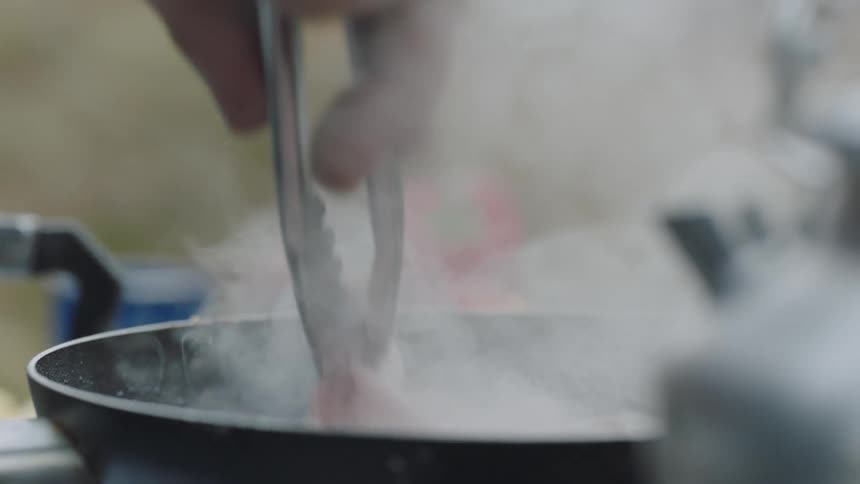 Person flips cooking bacon with tongs while smoke rises beside a blue pot