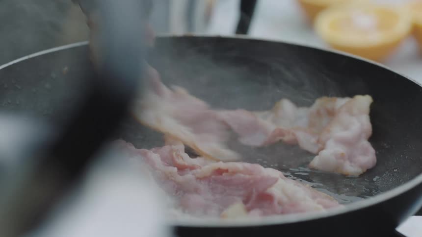 Person flips cooking bacon with tongs with oranges to the side
