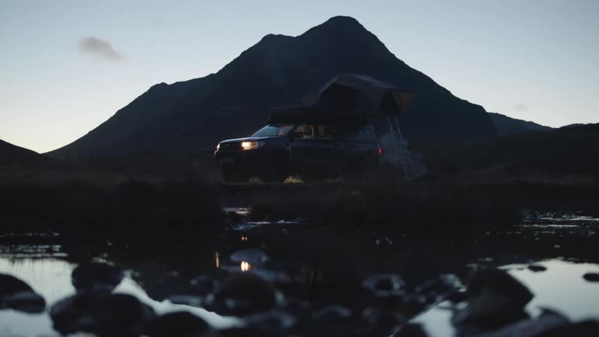 Light comes on in tent sitting partly on roof of vehicle near flowing lake and backed by mountain