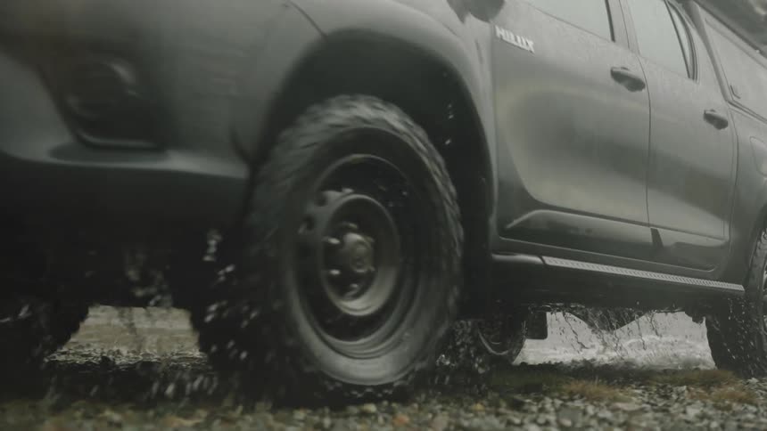 A vehicle drives through water on a gravel road near hills