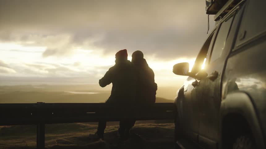 Couple sits on rail near packed vehicle looking over view as sun sets