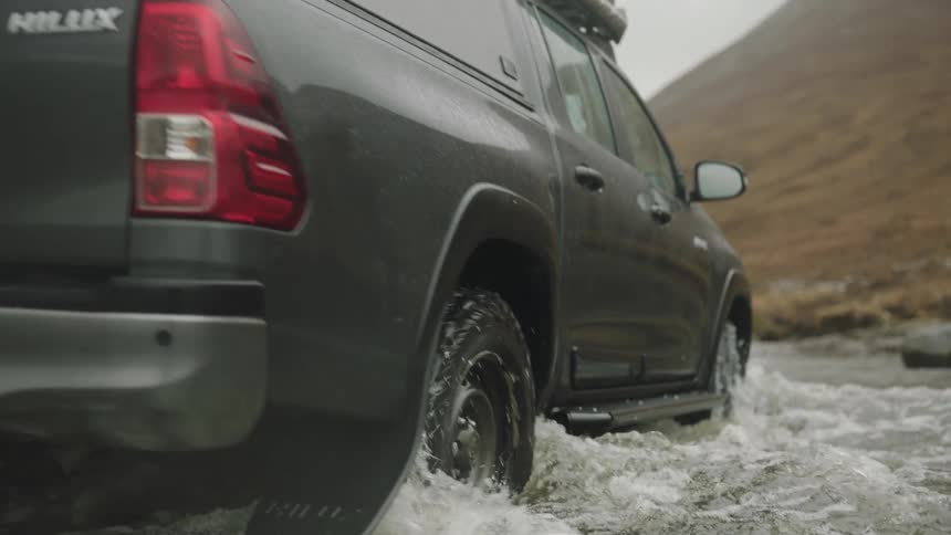 A truck drives through water near a creek bed and hills