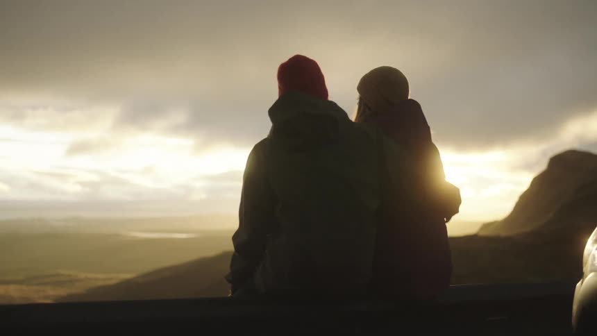 A couple sits at the top of a mountain top with a car parked behind them