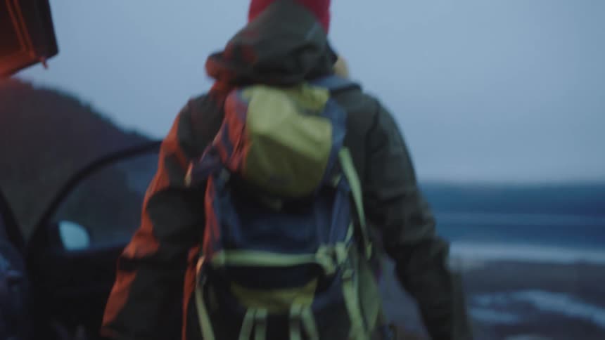A male hiker puts on his backpack before walking around a vehicle to greet a female hiker on a cloudy evening
