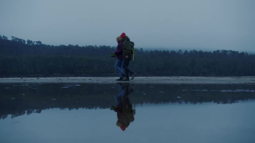 Two people with backpacks walk along the waters edge in the evening