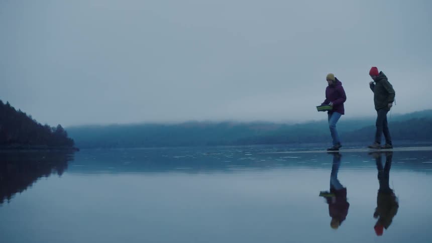 Two people walk out to the edge of a lake and crouch down near the water