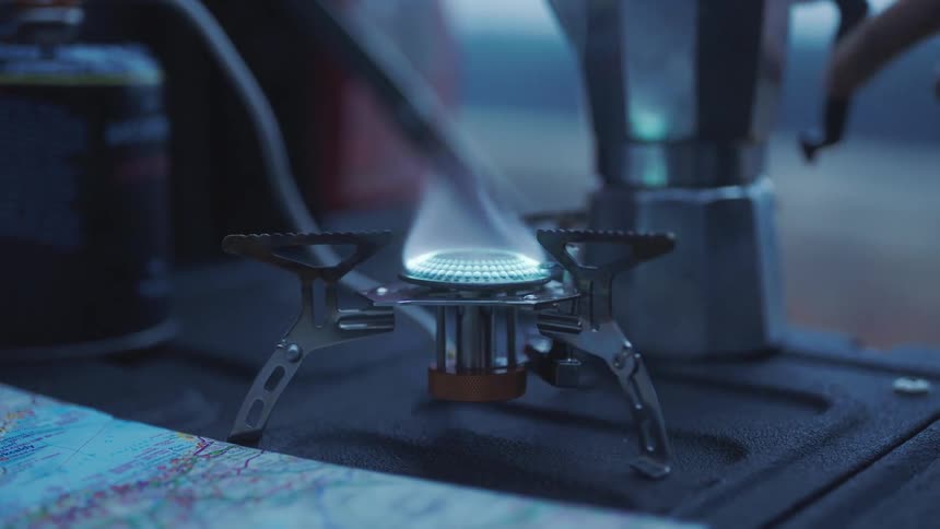 Person lights portable gas burner sitting on truck tailgate near map before placing coffee pot on burnier