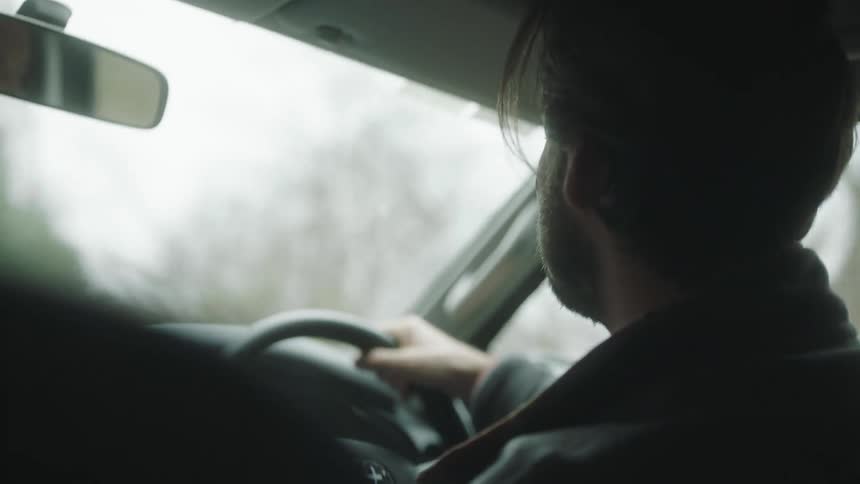 A man smiles and laughs as he drives a car and then a woman reaches over and rubs his hair