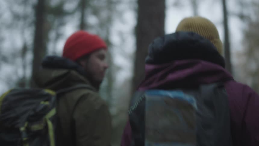A man retrieves a map from the backpack of a woman hiking beside him through a woodland on a cold day