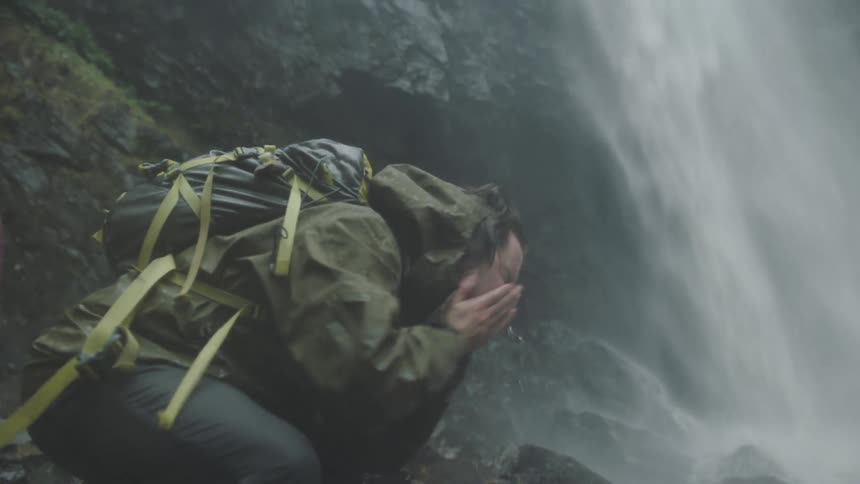 A man stands at the bottom of a waterfall scubs his face with the water and lifts his arm in celebration