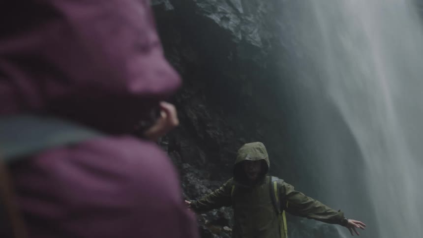 Man posing in front of a waterfall pouring down the side of a mountain while a person holding a camera takes pictures