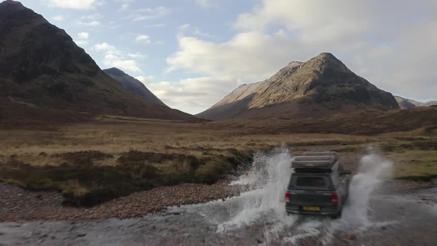 A vehicle drives across the grasslands and through a river towards mountains