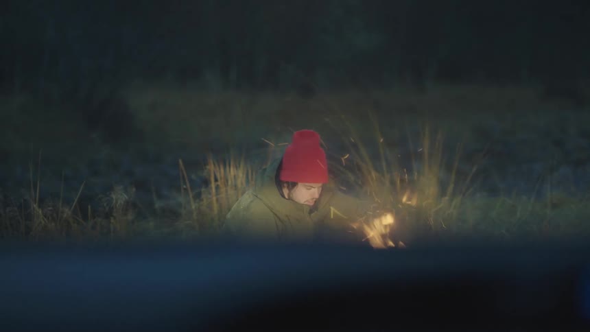 Man kneeling down in the evening in front of a parked car with windshield wipers on starting a campfire