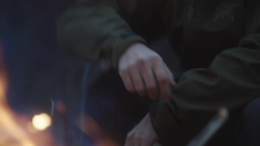 A man warms his hand sitting outside by a campfire
