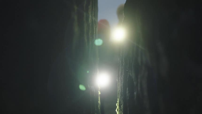 Man and a woman outdoors searching through a crack in a rock wall with flashlights